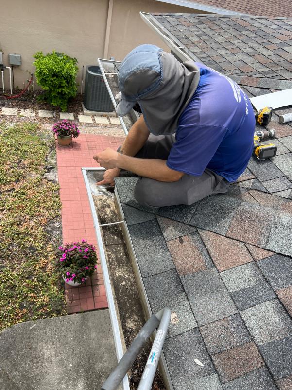 worker cleaning a gutter on a sloped roof using a tool with plants and a ladder nearby
