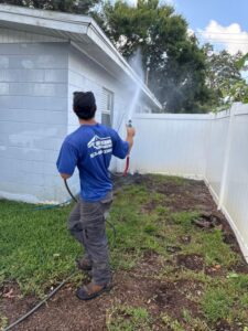 technician using a hose to clean the side of a house with washing spray in a grassy area
