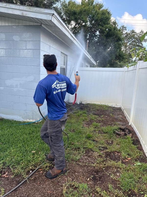 technician using a hose to clean the side of a house with washing spray in a grassy area