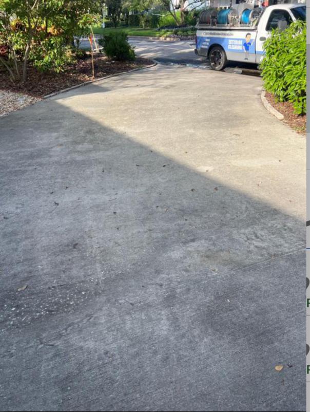 a concrete driveway showing signs of dirt and debris with a service vehicle parked nearby and lush greenery around