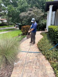 a worker using a pressure washer on a brick pathway surrounded by landscaping and greenery