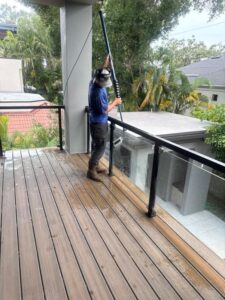 worker using a pole to clean a deck and glass railing in a residential area surrounded by greenery