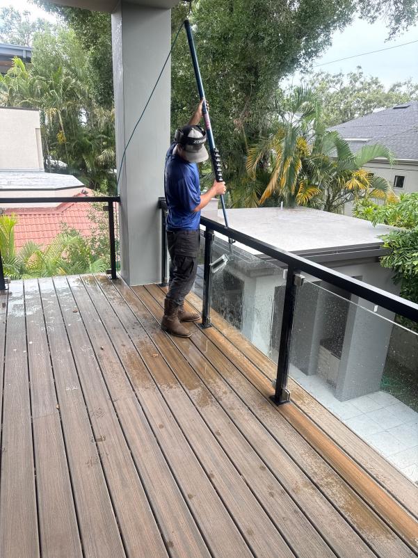 worker using a pole to clean a deck and glass railing in a residential area surrounded by greenery