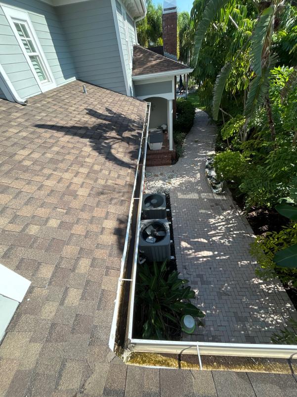 cleaning gutters with visible dirt and debris along the edge of a roof near air conditioning units and landscaping