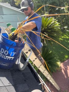 man clearing debris from a gutter using a bucket and gloves while on a ladder near a house