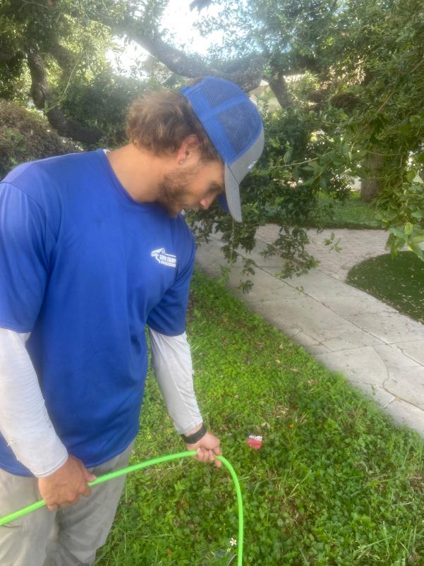 a worker assessing the garden area holding a green hose near overgrown grass and plants in a residential setting