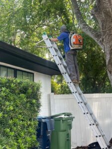 worker using a ladder to clean gutters on a house wearing a backpack blower with greenery in the background