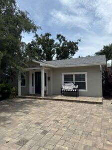gray house with a porch and a bench in front surrounded by trees and landscaping on a paved driveway