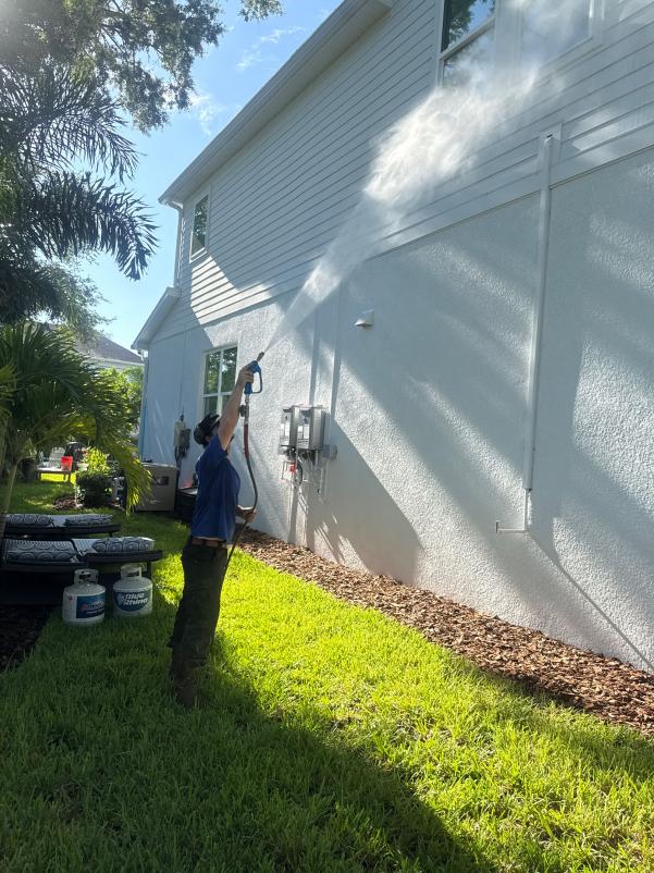a worker using a pressure washer on a house exterior with landscaping visible in the foreground and equipment nearby