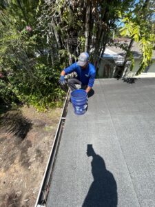 worker cleaning gutters on a roof using a bucket surrounded by trees and shrubbery under clear blue skies