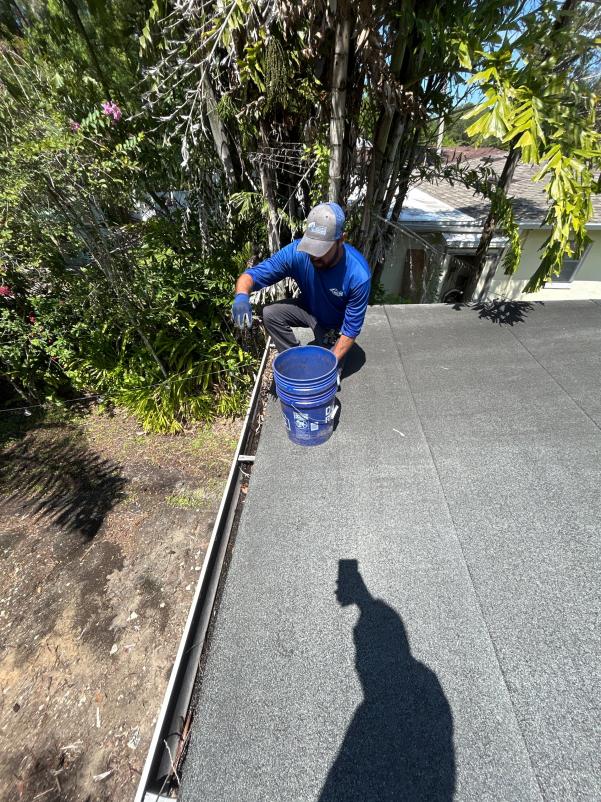 worker cleaning gutters on a roof using a bucket surrounded by trees and shrubbery under clear blue skies