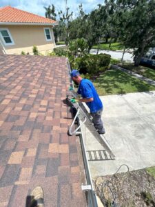man cleaning a roof using a ladder and equipment in a residential area with greenery and a driveway nearby