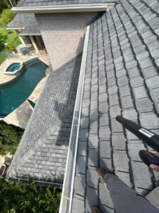 cleaning gutters on a sloped roof using a blower tool with a view of a pool and landscaping below