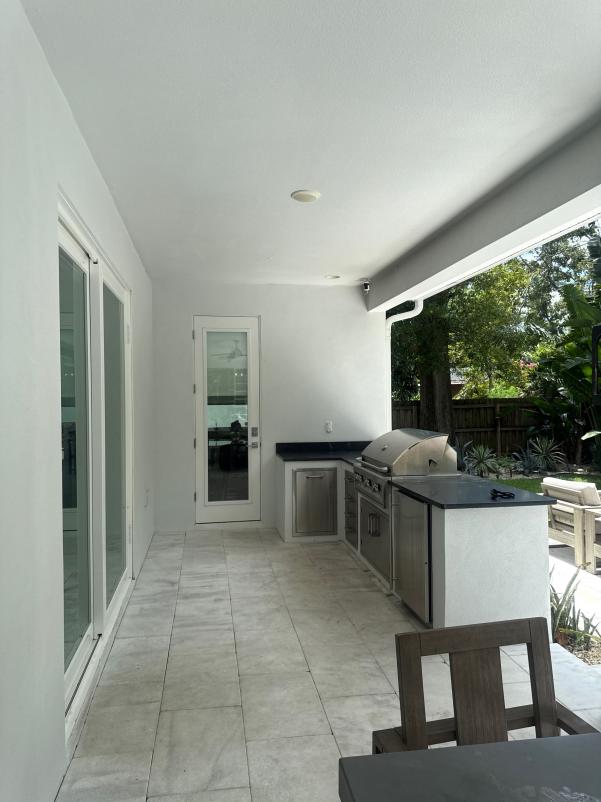 outdoor kitchen area with stainless steel grill and patio furniture on tiled flooring surrounded by greenery and a fence