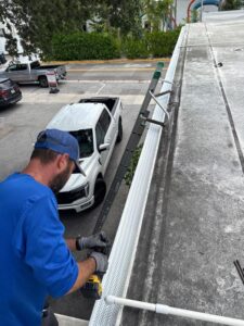 employee installing gutter system on a roof using a power drill with a ladder and parked truck below