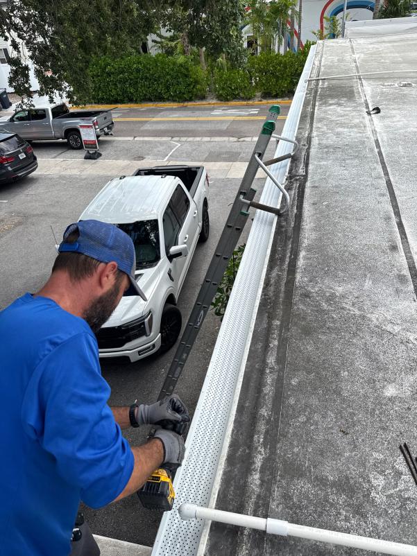 employee installing gutter system on a roof using a power drill with a ladder and parked truck below