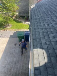 man cleaning gutter with gloves wearing a hat standing on driveway beside trash bins and a clear lawn