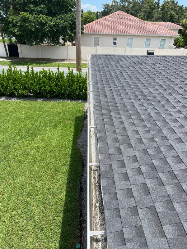 gutter cleaning near a gray shingled roof with visible debris accumulation and well-maintained green lawn in background